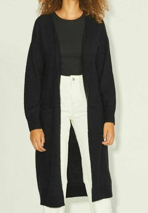 Personne portant un long cardigan noir ouvert sur un haut noir et un pantalon blanc taille haute, debout devant un fond uni.