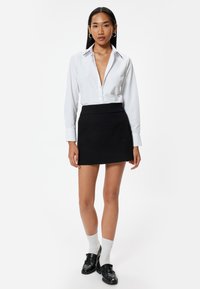 Blusa blanca de manga larga con escote en V profundo y un sutil detalle de cuello, combinada con una falda mini negra, calcetas blancas y mocasines negros.