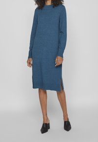 Kvinna iklädd en blå långärmad stickad klänning med sidoklyftor och svarta spetsiga blockklacksmules, stående mot en neutral bakgrund.