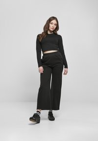 Schwarzes, langärmliges, kurzes Oberteil, kombiniert mit lockeren, schwarzen, hoch taillierten Hosen. Das Modell trägt schwarze Schuhe und schwarz-weiß gestreifte Socken.
