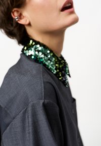 Zwarte shirt met een collier van pailletten in tinten groen en blauw. De stof heeft een gestructureerde uitstraling en de kraag is versierd met glanzende versieringen.