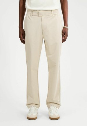 Persona che indossa pantaloni beige a gamba dritta con tasche laterali e scarpe da ginnastica bianche, in piedi davanti a uno sfondo bianco uniforme.