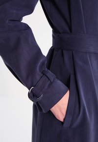 Námořnicky modrý trenčkot s páskem v pase, s hladkou texturou, detailovanými knoflíkovými manžetami a bočními kapsami.