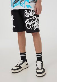 Pantaloni corti neri con disegni graffiti bianchi, abbinati a scarpe da ginnastica in bianco e nero e calze nere a righe. Stile streetwear casual.