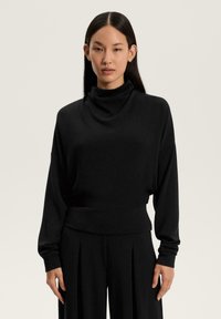 Tânără cu păr drept și negru, purtând un top lung negru cu mâneci lungi, cu guler înalt, și pantaloni largi negri, pe un fundal simplu.
