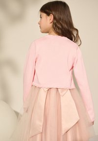 Cardigan rosa lavorato a maglia con orlo smerlato, abbinato a una gonna a balze in tulle con grandi fiocchi di raso in un delicato colore pesca.