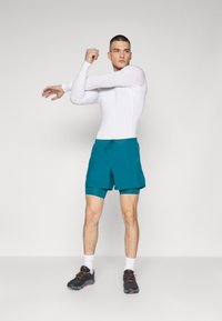 Langærmet hvid tætsiddende top med teal shorts. Shortsene har en snøre og et indvendigt lag, parret med sorte træningssko og hvide sokker.