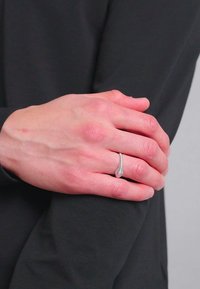 Srebrni prsten s uvijenim dizajnom, s teksturiranom površinom, nosi se na ruci koja miruje na tamnoj tkanini.