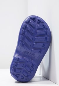 Suela de zapato de goma morada con una superficie texturizada, que presenta una banda de rodadura estampada y una etiqueta con el logo. Acabado suave con canales poco profundos y protuberancias.