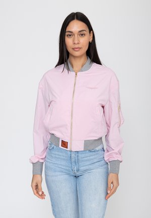 Frau mit langen dunklen Haaren, die eine hellrosa Bomberjacke mit grauen Bündchen und Kragen trägt, kombiniert mit hellblauen Jeans, stehend vor einem weißen Hintergrund.