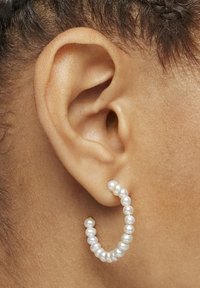 Zbliżenie ucha z kolczykiem kołowym z białych pereł na jasnobrązowej skórze i splecionych włosach.