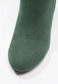 Botín de tobillo verde de ante con una textura suave, punta afilada y un brillo sutil. La suela es negra, contrastando con el material superior.
