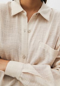 Femme portant une chemise beige texturée à boutons, avec un col et une poche poitrine, les bras croisés à la taille.