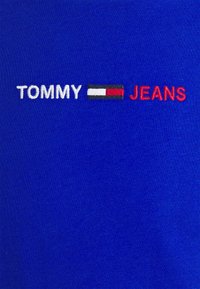 Modrá tkanina s vyšitým nápisem "TOMMY" bílou barvou, obdélníkovým logem v červené, bílé a černé barvě a nápisem "JEANS" červenými písmeny.