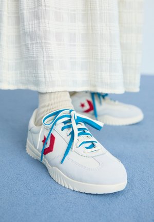 Baskets blanches avec des accents rouges et bleus, en tissu texturé, lacets plats, semelles en caoutchouc et détail de logo sur le côté. Portées avec des chaussettes de couleur claire.