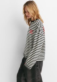 Jong persoon met schouderlang blond haar, gekleed in een zwart-wit gestreept shirt met lange mouwen en rode tekst, en donkere, losse broek, kijkt naar beneden.