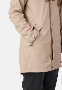 Veste imperméable beige dotée d'une fermeture à boutons-pression, de poches latérales et d'une texture lisse, avec des coutures visibles et une coupe décontractée.