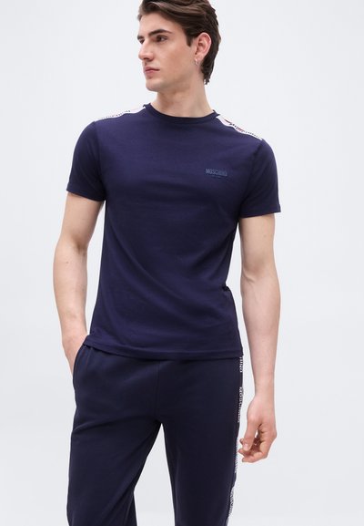 Homme portant un t-shirt Moschino bleu marine et un pantalon assorti avec des bandes à logo, debout les mains dans les poches, regardant sur le côté.