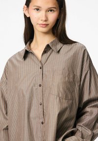 Rjava črtasta srajca s ovratnikom in gumbnim zapenjanjem spredaj, ki ima žep na levi strani prsnega dela in sproščen kroj, izdelana iz lahkega materiala.