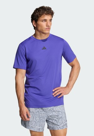 Hombre con camiseta morada de Adidas y pantalones cortos grises con estampado, de pie con una mano en la cadera, mirando hacia un lado con un fondo liso.