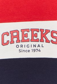 Tessuto a strisce rosso, bianco e blu navale; presenta il testo "CREEKS ORIGINAL Dal 1974" in lettere grasse rosse e nere.