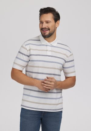 Uśmiechnięty mężczyzna z brodą, ubrany w białą koszulkę polo z beżowymi i niebieskimi poziomymi paskami oraz niebieskie dżinsy, stojący na jasnym tle.