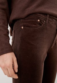 Pantalones de pana marrón con un corte ajustado, que presentan cordones sutiles en la tela, remaches de cobre y bolsillos en la parte delantera y los laterales.