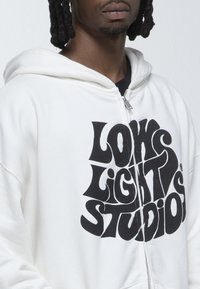 Felpa con zip bianca caratterizzata da un grande e audace design testuale grafico nero che recita "LOW LIGHT STUDIOS". Tessuto morbido, vestibilità comoda, senza motivi visibili.