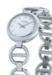 Montre-bracelet en argent avec un cadran rond blanc, des index en points, et un bracelet à maillons semblables à une chaîne, orné d'accents en cristal et d'une finition métallique lisse.