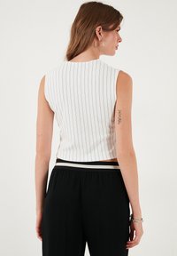 Kratka bela majica z črnimi vertikalnimi črtami, brez rokavov, z rebrasto teksturo, v kombinaciji s črnimi hlačami z črtastim pasom.