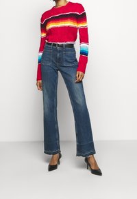 Pull rouge côtelé avec des rayures multicolores, associé à un jean bleu taille haute à jambes larges et des escarpins noirs à bouts pointus.
