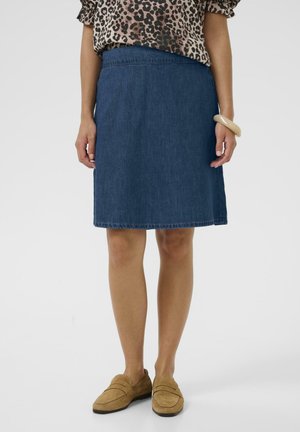 Kvinde iført en knælang blå denimnederdel, leopardprintet top, beige armbånd og lysebrune loafers, stående på en hvid baggrund.