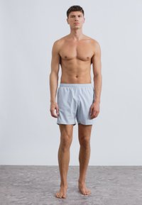 Ανοιχτό μπλε μαγιό σορτς από ύφασμα γρήγορου στεγνώματος, με ελαστική μέση και πλαϊνές τσέπες, φορεμένο από μοντέλο που στέκεται σε υφασμένη επιφάνεια.