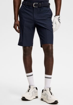 Man draagt marineblauwe korte broek, witte riem, witte golfhandschoen aan rechterhand, witte sokken met zwarte strepen en witte sneakers met zwarte details.