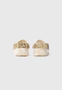 Sabots en suède beige avec des boucles décoratives, des accents de perles et de strass sur la sangle, une semelle en caoutchouc texturée et une base à motifs.
