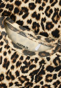 Overdel i leopardmønstret stoff med ribbet halslinning med sorte og hvite striper. Bakgrunnen er lys beige med brune og svarte flekker.