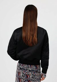 Mustat satiin bomber-takit, joissa on ribattu hihansuut ja helma, ja jotka ovat silkkisiä ja kerroksellisia hihoissa. Päällä kuvioidut housut.