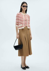 Pruhovaný krémový a červený cardigan přes hnědou skládací sukni, doplněný černou kabelkou a černými lodičkami. Modelka nosí sluneční brýle.