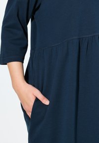 Tummansininen, polven mittainen mekko pehmeästä kankaasta, jossa on kolmivartiset hihat, vyötärölle kerääminen ja sivutaskut.