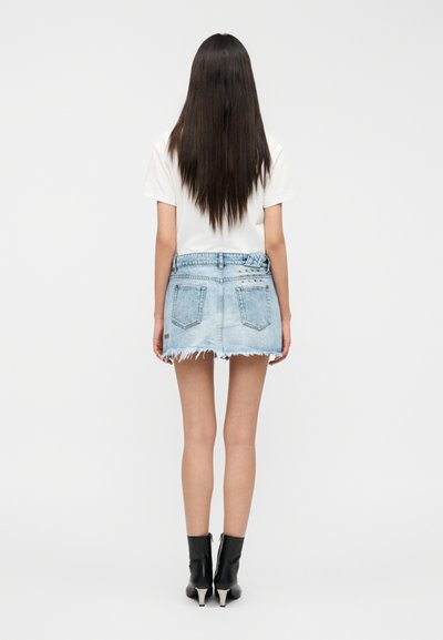 Μίνι denim φούστα σε ανοιχτό μπλε με ξεφτισμένο τελείωμα, συνδυασμένη με λευκό T-shirt και μαύρα μποτάκια αστραγάλου. Διαθέτει δύο πίσω τσέπες και ορατές ραφές.