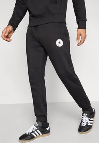 Black sweatpants with a fitted design, elastic cuffs, and a round patch detail. Worn with black and white sneakers featuring three stripes.