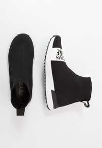 Svarta tennisstrumpor i ankellängd tillverkade av stickat material, med ett vitt logoband och en strukturerad gummisula med greppdesign.