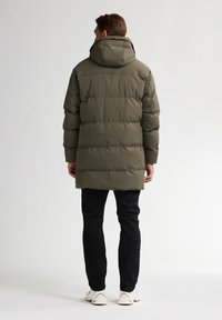 Manteau déperlant vert olive avec capuche, présentant un design matelassé, des manches longues et une coupe mi-longue. Associé à un pantalon noir et des chaussures blanches.
