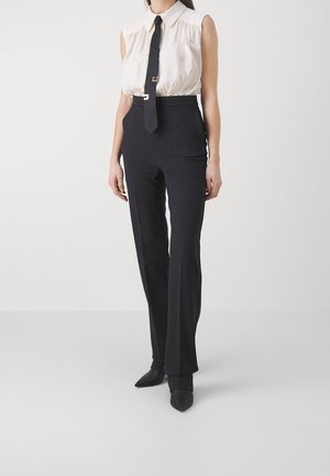 Kvinde iført ærmeløs hvid bluse, sort slips, sorte bukser med høj talje og sorte spidse sko mod ensfarvet baggrund.