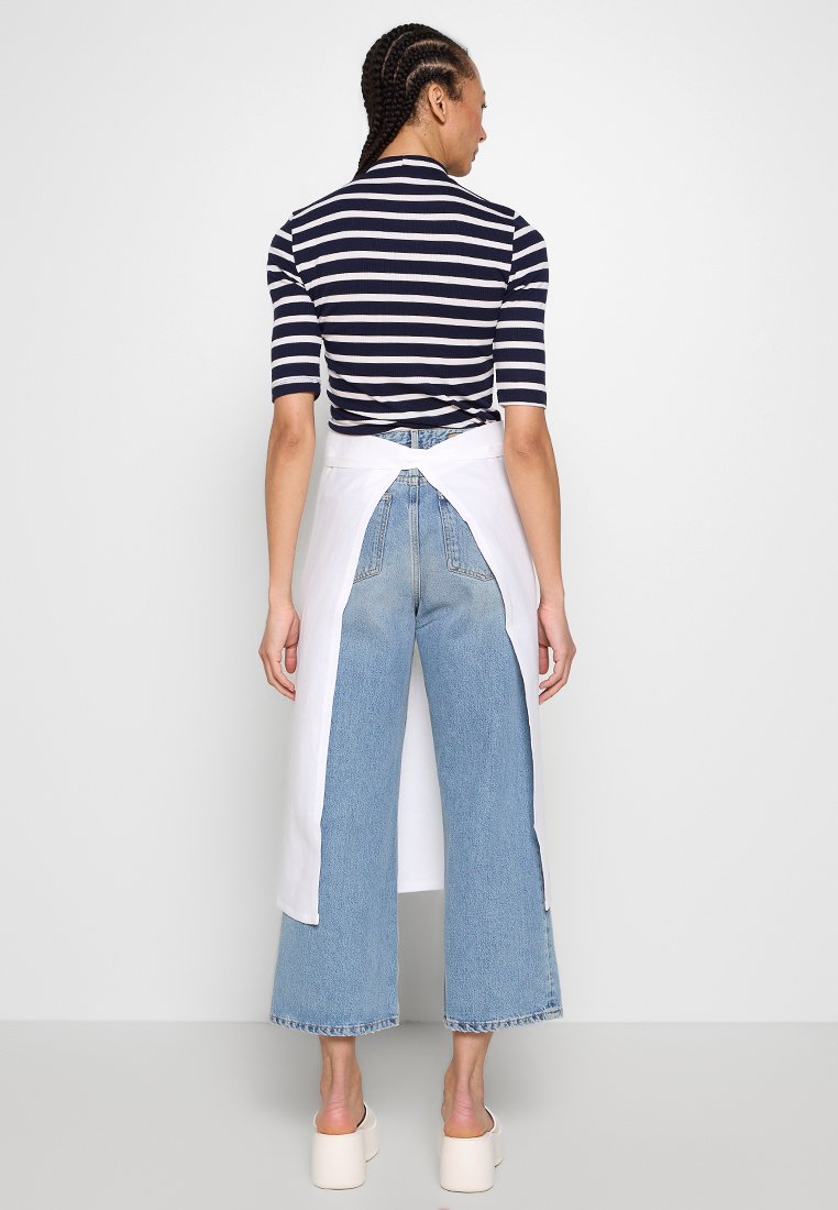 Top a righe blu navy e bianche con maniche corte, abbinato a jeans svasati blu chiaro. Sovrapposizione a grembiule bianca, legata sul retro, che enfatizza la semplicità.