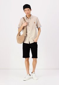 Camisa de manga corta beige con puños azul claro, bolsillo en el pecho, pantalones cortos negros y zapatillas blancas. Lleva un bolso de hombro en color marrón.