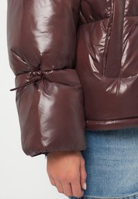 Hnedá, lesklá puffer bunda s detailom zhrnutého rukáva, sťahovanie pomocou šnúrky. Detailný záber na manžetu a ruku na denim.