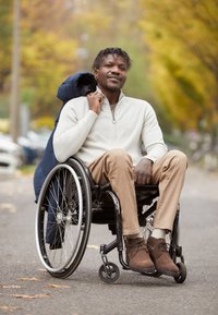 Muž v bílém zapínacím svetru a béžových kalhotách sedí v černém vozíku s šedými koly, má na sobě hnědé semišové boty, přes ramena má přehozený modrý kabát.