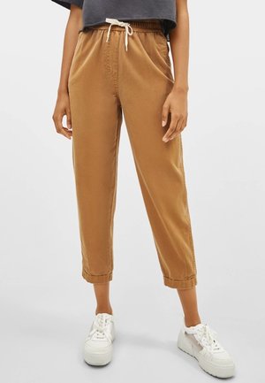 Vrouw draagt cropped bruine broek met trekkoord en omgeslagen pijpen, gecombineerd met witte sneakers, staand tegen een effen achtergrond.