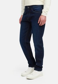 Dark blue denim jeans with a slim fit, featuring front pockets, a zip fly, and contrast stitching, paired with white sneakers.
